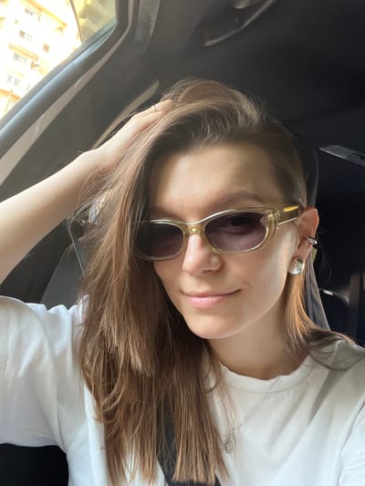A young woman with long, light brown hair and fair skin is captured in a close-up, selfie-style shot. She is wearing sunglasses with clear, tinted lenses and a thin, silver necklace. Her left hand is resting on her head, near her hair. She has a subtle smile on her face. The background appears to be the interior of a car, with dark upholstery visible. Through the car window on the left, a bright, sunlit scene with buildings is visible, suggesting it is daytime. The lighting indicates it is a sunny day. The woman is wearing a white t-shirt. The image has a relaxed and casual mood.