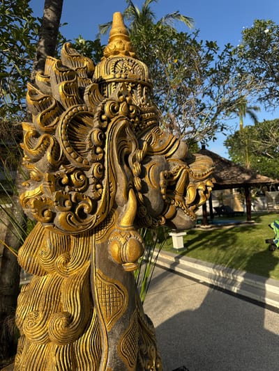 The image features a close-up of a heavily ornamented, gold-accented stone sculpture situated outdoors in Jimbaran, Indonesia. The sculpture's intricate details suggest a traditional Balinese style, with elaborate carvings and a weathered appearance. The sculpture is positioned in front of a paved area and a well-manicured lawn. A partially visible building with a thatched roof is visible in the background. The scene is captured under clear, bright daylight.