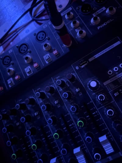 The image shows a close-up, overhead view of audio mixing equipment, likely a soundboard and a separate smaller mixer, illuminated by blue lighting. The larger mixer in the foreground features multiple channels with knobs, faders, and indicator lights in green and blue. Labels visible include "PEAK," "GAIN," "PAN," "MID," "LOW CUT," "FREQ," "FX LEVEL," and "FX AUX." The smaller mixer in the background has XLR and 1/4-inch input/output jacks, labeled "MIC/LINE," "PHONES," and "C.ROOM." It also shows an "MP3 PLAYER RECORDER" section with a USB port. Cables are visible connected to the equipment. The overall environment appears to be indoors, possibly a music venue or studio, though the city of Isola delle Femmine, Italy is not discernible from the visual information provided. There are no people present in the image.