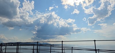 This is a wide shot taken from a railing overlooking a body of water. The sky is filled with dramatic clouds, with sunlight breaking through in places, creating visible sunbeams. The water is calm, reflecting the sky. In the distance, a low-lying landscape with hills and trees is visible, suggesting a natural setting. The railing in the foreground is dark and metallic, with a simple design. The scene appears to be a peaceful, scenic view, possibly from a promenade or a pier. The location is identified as Lochau, Austria.