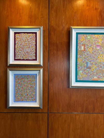 Three framed paintings are displayed on a wooden wall with vertical seams. The top painting is larger than the two below, which are placed side-by-side. The top painting has a gold frame with a white and teal inner border, and the painting itself features a mosaic-like pattern with squares and rectangles of various colors. The bottom left painting is framed in gold and white with a dark purple inner border, and displays an abstract pattern with dots and organic shapes in shades of orange, yellow, and white. The bottom right painting has a gold and white frame with a light blue border and a mosaic style painting with squares and rectangles in various colors. The wooden wall appears to be indoors and well-lit. The location is not discernible from the image, therefore no location can be derived.