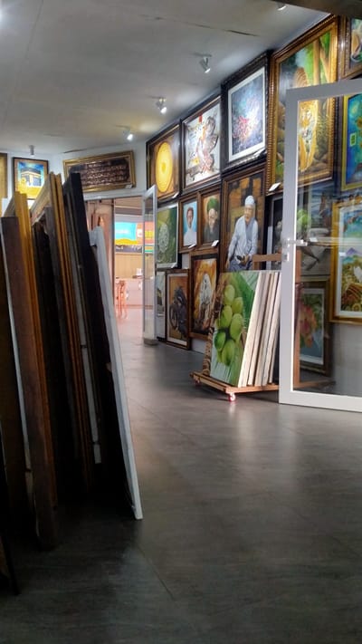 This is a full, eye-level shot of an indoor art gallery, likely in Nyamat, Indonesia, featuring numerous framed paintings displayed on walls and leaning against each other. In the foreground, on the left, is a collection of art frames stacked vertically, leaning against a wall. The gallery floor is a polished concrete.
The main focus is the art on display. The walls are covered with paintings of various sizes and themes. One prominent large painting shows a tiger, framed in dark wood. To its right, a painting of a tiger walking through trees is visible. Further to the right, a tall white framed painting shows a landscape with a body of water. Another large framed piece to the left of the tiger painting depicts what appears to be a religious scene with figures.
Below these, a variety of smaller framed artworks are visible. There's a painting of a motorcycle, a portrait of a man in a green turban, and another portrait of an older man. To the right, a painting of green coconuts on a branch is propped up on a wheeled easel.
In the background, through an open doorway, a brightly lit area is visible with chairs and tables, suggesting a seating area or a cafe within the gallery. The ceiling has multiple track lights illuminating the artwork. The overall impression is of a well-stocked and professionally displayed art collection.