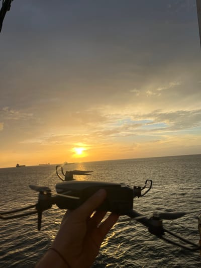The image displays an expansive outdoor scene captured at either sunrise or sunset over a large body of water. The composition is horizontally oriented, featuring a vast sky above and water below, illuminated by a warm, golden light.
In the foreground, towards the lower right of the frame, a human hand is visible holding a dark-colored drone. The drone, equipped with propellers, is silhouetted against the bright backdrop of the sky and the reflective water, suggesting it is being prepared for flight or has just landed.
The sky gradient moves from vibrant golden and orange tones near the sun to softer blues and grays higher up. Scattered clouds are present, catching the warm light and adding texture to the heavens. The sun is a bright, intense orb positioned low on the horizon, creating a prominent lens flare and casting a brilliant glow across the scene.
The water surface appears calm with gentle ripples, reflecting the golden light of the sun. This reflection creates a shimmering effect that extends across the water towards the foreground. In the distant background, along the horizon line, several dark silhouettes of ships or boats are visible, indicating maritime activity.
The time of day is clearly either sunrise or sunset due to the low angle and intense warmth of the sun's light. The weather appears clear with some cloud cover. No discernible text is visible within the image. The scene conveys a sense of tranquility and the natural beauty of the golden hour over open waters.