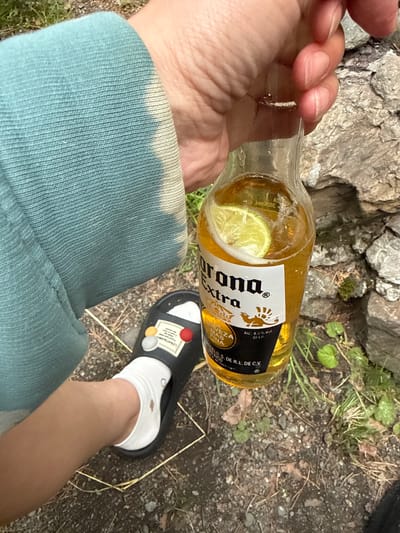 An individual's hand, with visible fingernails, is holding a clear glass bottle of Corona Extra beer. The bottle contains a light-colored liquid and a slice of lime is visible floating inside. The bottle has a white label with the "Corona Extra" logo. The individual is wearing a teal-colored long-sleeved garment and a white ankle sock. A black slide sandal with colorful decorative elements is on the individual's foot. The scene is outdoors, with a rough, rocky surface and sparse green vegetation in the background. The location is identified as Cheposh, Russia.
