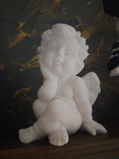 A white porcelain figurine of a cherub is positioned on a dark wooden surface in front of a textured dark green and gold background. The cherub is depicted in a seated position, with its head resting on its right hand and its legs crossed. It has small wings on its back. The lighting is dim, casting subtle shadows. The scene appears to be an indoor setting. There is no information about the location being in Сухарево, Russia.