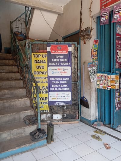 The image shows the exterior of a storefront located in Katapang, Indonesia (latitude: -6.99, longitude: 107.552). The storefront has a blue door with a metal grate on the front. To the left of the door, there is a set of stairs leading up to a higher level. On the side of the building, there are two yellow signs advertising services such as "Top Up Dana OVO GOPEY" and "Top Up Tarik Tunai Semua E-Money." A broom is propped up against the wall, and there are three pairs of shoes on the floor near the bottom of the stairs. Two pairs of flip-flops and one pair of rainboots are visible. The stairs are made of concrete and appear to be worn. The storefront has a white tiled floor and the walls are painted white. There are several objects on display on the wall behind the blue door. There are no people in the image.