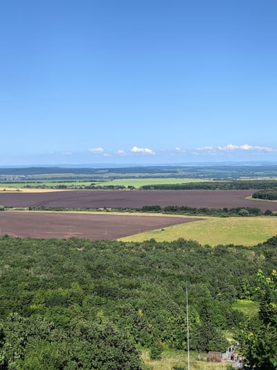 A wide, panoramic view of a rural landscape in Akhmerovo, Russia, under a bright blue sky with sparse white clouds. The foreground is dominated by a dense, lush green forest. Beyond the forest, several agricultural fields are visible. The closest fields are a dark brown, recently plowed earth. Further out, there are fields of light green and yellow crops. In the distance, rolling hills and a hazy horizon suggest the presence of more fields and possibly a distant body of water or low-lying land. The overall impression is one of vast, open spaces and agricultural activity.