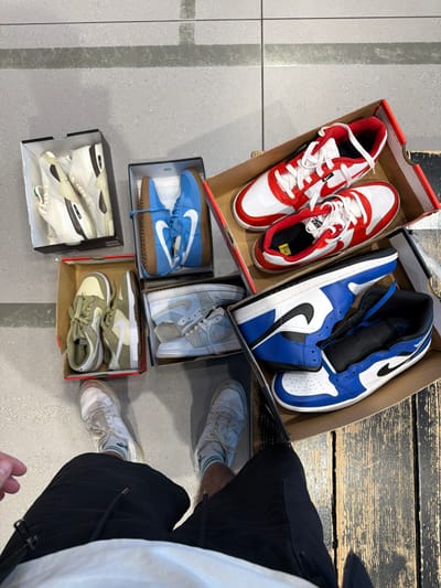 An overhead photograph depicts the lower body of an individual standing in a retail environment in Rishon LeZion, Israel, surrounded by multiple open shoe boxes containing athletic footwear. The person is wearing black shorts, white socks with green stripes, and a pair of white sneakers. On the floor, six open shoe boxes are arranged around the person's feet. These boxes contain various Nike brand sneakers: a pair of cream/off-white chunky trainers, a pair of olive green low-top sneakers, light blue Air Force 1s with a gum sole, red and white low-top sneakers (one with a yellow size sticker), light grey/white textured sneakers, and blue, white, and black Air Jordan 1 high-top sneakers. The flooring transitions from light grey square tiles on the left to dark, distressed wooden planks on the right. The scene suggests a shopping activity in a shoe store.