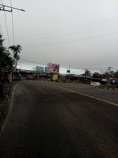 This image depicts a street scene on an overcast day, reportedly located in the city of Deothang, Bhutan. However, visible text on billboards, such as "SEGER ALAM MANA RAKA BANS" and "Love Style" alongside an advertisement featuring young women, suggests an Indonesian linguistic context.
The scene features a damp, dark asphalt road with a double yellow line visible in the center-right. Overhead, numerous black utility wires crisscross against a uniformly grey, cloudy sky. A street lamp on a pole extends from the upper left.
On the left side, the road curves slightly uphill, flanked by a bank covered in green vegetation, including small trees and bushes. A row of small, single-story commercial buildings or stalls, exhibiting various colored facades and corrugated metal roofs, lines this curving road. A white circular regulatory sign with a red border is affixed to a pole near these structures. A yellow compact commercial vehicle, possibly a minivan or small truck, is parked among these buildings. One person is partially visible standing near this yellow vehicle, and another person can be discerned further down the road on the left.
In the mid-ground, a central median or island, adorned with small potted plants, separates sections of the road or an intersection. Beyond this median, two large billboards dominate the view. The left billboard displays white text, partially reading "SEGER ALAM MANA RAKA BANS," on a blue background. The right billboard features a pink background with a "Love Style" logo and an image of several young women, appearing to be an advertisement for a beauty or fashion product. Further behind these billboards, more buildings with varied construction and utility poles are visible. A white passenger van is parked on the far right side of the road, facing right. In the far background on the right, a darker, forested area is discernible beneath the overcast sky. No direct interactions between people are evident.