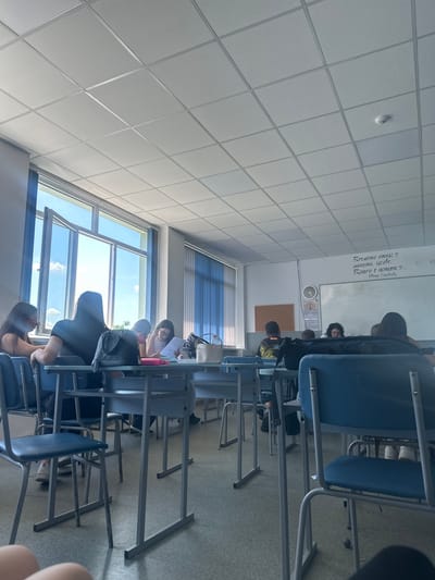 The image depicts the interior of a classroom in Targovishte, Bulgaria. Several young people, predominantly female, are seated at desks. The classroom is equipped with standard school furniture, including metal-framed desks and blue-upholstered chairs. Large windows, some of which are open, are visible, allowing natural light to enter. The ceiling is made of acoustic tiles, and a smoke detector is present. The walls are plain, with one wall featuring a whiteboard and a clock. There is some writing on the whiteboard, but it is illegible at this resolution. The floor is covered in a light-colored carpet. The overall atmosphere appears to be one of a school setting during a lesson or study period.
