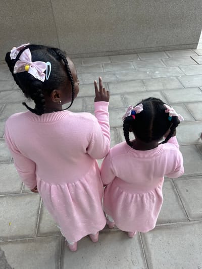 Two young Black girls are standing on a paved walkway. They are wearing matching pink dresses and have braids in their hair adorned with bows. The girls are captured from an overhead perspective. One girl is extending her fingers. The surrounding environment consists of gray, rectangular paving stones. The location is likely within the city of Sant Vicent del Raspeig, Spain.