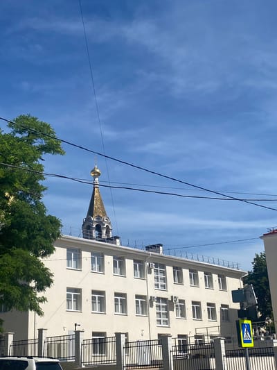 The image shows a bright, sunny day in a city setting, possibly Sevastopol, Ukraine. The perspective is looking upwards towards a clear blue sky with scattered, wispy clouds. Prominently featured are several black electrical wires crisscrossing the frame.

To the right, a tall, modern, light-colored building with numerous windows dominates the view. The building appears to have several floors, with some windows showing closed blinds or curtains. Air conditioning units are visible mounted on the exterior walls. Balconies with black railings are also present on some levels. A pedestrian crossing sign, a yellow square with a blue symbol of a pedestrian, is mounted on a pole next to the building.

In the middle ground, peeking above the trees and electrical wires, is the golden dome of a church or religious building. The dome is intricately detailed and topped with a small spire, suggesting architectural significance.

In the foreground, lush green trees with dense foliage are visible, partially obscuring the lower parts of the building and the church dome. The overall impression is one of an urban environment with both modern infrastructure and historical or religious elements, bathed in bright daylight.