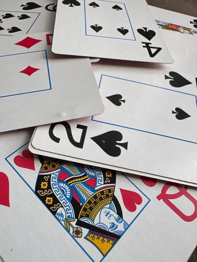 A close-up shot shows several playing cards scattered randomly. Prominent cards include the Four of Spades, Two of Spades, and Queen of Hearts. Partial views of other cards displaying diamond and spade suits are visible, partially obscured. The cards overlap each other, creating a chaotic arrangement with a mix of white backgrounds and suit symbols.