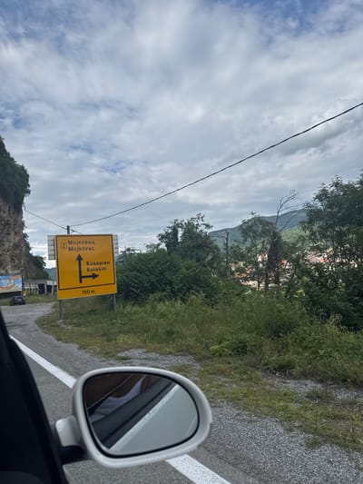 The image shows a road sign indicating directions to Mojkovac and Kolasin, in Babljak, Montenegro. The sign is yellow with black text and arrows. One arrow points straight up for Mojkovac, and another points right for Kolasin. Below the destinations, "150 m" is visible. The sign is located on the side of a paved road, with a rocky cliff face and vegetation on the left. In the background, there are trees and hills, with some buildings visible through the foliage. A white car's side mirror is in the foreground, reflecting a person wearing a white tank top. The sky is cloudy.