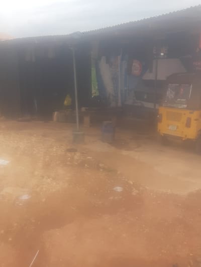 The image captures a dimly lit outdoor scene, likely on a dusty road in Enugu, Nigeria. The foreground is dominated by reddish-brown dirt and scattered stones. In the background, a structure with a corrugated metal roof appears to be a shop or market stall. A yellow auto-rickshaw, a common mode of transport in Nigeria, is parked to the right. The rickshaw has a banner on its rear that reads "THE RETURN" and "OUT NOW!". Behind the rickshaw, a large poster with text is visible on the wall of the structure. To the left, a dark interior of the structure is seen, with a yellow plastic bag hanging and some unidentifiable objects. The overall impression is one of a utilitarian, possibly commercial, area in a developing urban environment, with limited visibility due to the lighting conditions and potential dust in the air.