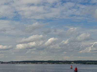 The image depicts a wide shot of a body of water, likely a river or lake, with a distant shoreline. The sky is partly cloudy with cumulus formations against a blue backdrop. On the shoreline, a strip of trees forms a dark green band, behind which various structures are visible, including buildings and what appear to be construction cranes. The foreground of the water features two buoys, one red with white markings and another primarily red. The overall scene suggests a moderately populated area with ongoing development, viewed from a distance across the water. The location is identified as Verhnij Uslon, Russia.