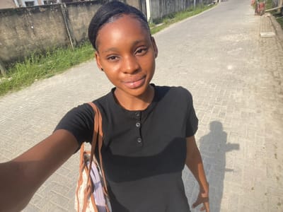 A young female, identified as being in Iranla, Nigeria, is depicted taking a selfie outdoors. She has dark skin and her dark hair is pulled back from her face. She wears a black, short-sleeved henley-style shirt with a buttoned placket. A light-colored shoulder bag with brown straps and a patterned design is visible on her left side. Her right arm is extended, holding the camera. The background shows a paved road constructed with light-colored rectangular interlocking pavers, stretching into the distance. To the immediate left of the subject, a weathered grey concrete wall is partially obscured by green grass and weeds. To the right, a narrower paved path runs parallel to the road, with a red and brown cart or trolley visible further down the street. The scene is brightly illuminated by sunlight, casting distinct shadows of the subject onto the paved surface to her right.