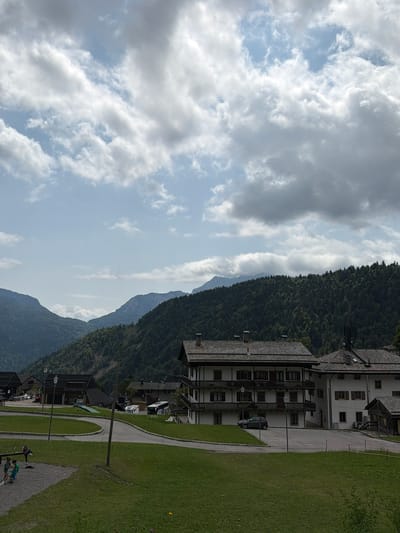 This is a medium-shot, eye-level, outdoor photograph of a village in Sauris / Zahre, Italy. In the foreground, there is a children's playground with a seesaw and a small cluster of people. A paved road curves through the scene, with a few buildings visible in the middle ground, including a multi-story building with balconies and a darker-roofed structure next to it. In the background, a densely wooded mountain slopes upward, with more distant, hazy mountains visible behind it. The sky is overcast with dramatic clouds. There are several vehicles in the scene, including a white campervan and a dark bus. A single wooden utility pole stands in the middle of the image. The overall impression is of a peaceful, rural mountain village.