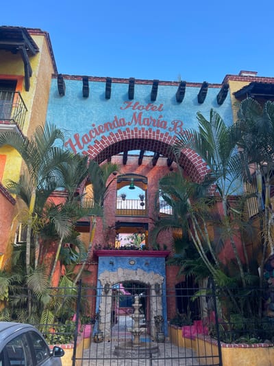 The image captures the exterior of the "Hacienda Maria B" hotel in Playa del Carmen, Mexico. The building is a vibrant mix of colors, predominantly yellow, orange, light blue, and red, and is adorned with palm trees. The name of the hotel, "Hacienda Maria B," is prominently displayed in red lettering. The building's architecture features brickwork around archways and windows. A wrought-iron fence partially obscures the ground level. A portion of a silver car is visible on the right side of the frame. The sky above the building is a clear blue.