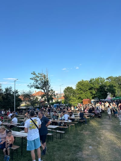 The image depicts an outdoor gathering of people, likely at a festival or event, in the city of Gorna Oryahovitsa, Bulgaria. The scene is characterized by numerous long wooden tables and benches set up on a grassy area, with many individuals seated and standing around them. The background reveals trees, buildings with tiled roofs, and a clear blue sky. People of various ages are present, engaged in conversation and enjoying the atmosphere. Some are holding drinks, and there are signs and tents visible in the distance, suggesting food and drink vendors. The overall impression is one of a community event taking place on a pleasant day.