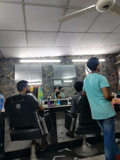 The image depicts the interior of a barbershop located at 23.784° N, 90.377° E in Dhaka, Bangladesh. The scene shows four men, three seated and one standing. The seated men are getting haircuts. The standing man is a barber, waiting to serve another customer. The barber is wearing a turquoise t-shirt and blue jeans. The men are seated in black leather barber chairs facing a mirror with a long, narrow, white-light fixture above. The walls are decorated with a black-and-gold marble-patterned wallpaper. There are three rectangular mirrors on the wall behind the chairs. One of the mirrors reflects an air conditioner unit on the wall behind the barber chairs. The floor is covered with a white tile, and there are some pieces of paper on the floor. The ceiling is white and has a white ceiling fan with a silver fan blade. The space is well-lit.