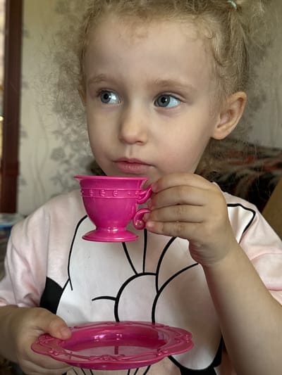 A young child, approximately 3-5 years old, with fair skin, light blonde, tightly curled hair, and prominent blue eyes, is depicted in a close-up, chest-up portrait. The child's gaze is directed towards the upper left. A small, light blue-green hair tie is visible in the hair above the right ear. The child wears a light pink long-sleeved top featuring a large, stylized black outline of a cartoon character (resembling Mickey Mouse) against a white oval on the chest. The right hand holds a bright pink plastic teacup with a decorative raised band and a small handle, positioned near the child's slightly parted lips, suggesting an act of drinking or play. The left hand holds a matching bright pink plastic saucer with an ornate, embossed rim. The background is an indistinct indoor residential setting in Nizhnyaya Gar', Russia, characterized by light-colored wallpaper with a subtle pattern and a dark, vertical element on the far left, possibly a door frame.