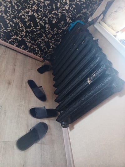 A low-angle, interior shot shows a dark, ribbed radiator taking up the right side of the frame. To the left of the radiator, on a light wood-grain floor, are three pairs of dark-colored slippers. One pair is lying on its side, partially obscured by another. The background features a dark wall with a beige ornate pattern and a light-colored baseboard. The scene is likely from a residential setting in Smolensk, Russia, judging by the style of the radiator and the commonality of such patterns in older Russian homes.