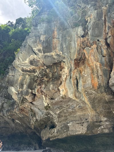 The image shows a cliff face with varied textures and colors, located in Ao Nang, Thailand. The cliff is composed of grey rock with streaks of orange and white, suggesting mineral deposits or water staining. Vegetation, including trees and vines, grows on the top and sides of the cliff. The lower part of the image reveals a sandy beach and a body of water. In the bottom left corner, a person is partially visible, standing on the beach. The lighting suggests it is daytime, with sunlight casting shadows and creating highlights on the rock surface. The overall scene depicts a natural coastal landscape.