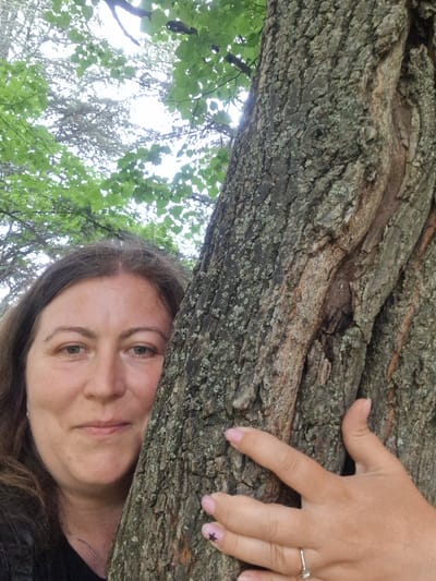 This is a close-up, low-angle shot of a woman resting her head on a large tree branch. Her eyes are open, and she appears to be looking towards the camera with a calm expression. Her hair is brown and falls around her head. A thin silver necklace is visible around her neck. Her left hand is resting on the tree branch, with her fingers spread slightly. She is wearing a silver ring on her left ring finger and has light pink nail polish on her fingernails, with a small black star design on one nail. The tree bark is rough and textured, with patches of green lichen. Above the branch, lush green leaves and branches are visible against a bright, overcast sky, suggesting an outdoor setting in a natural environment. The overall mood is peaceful and relaxed. The scene likely takes place during the daytime, given the brightness of the sky. While specific location cues are not overtly present in the image itself, it is stated to be located in Pancharevo, Bulgaria. No text is visible in the image.