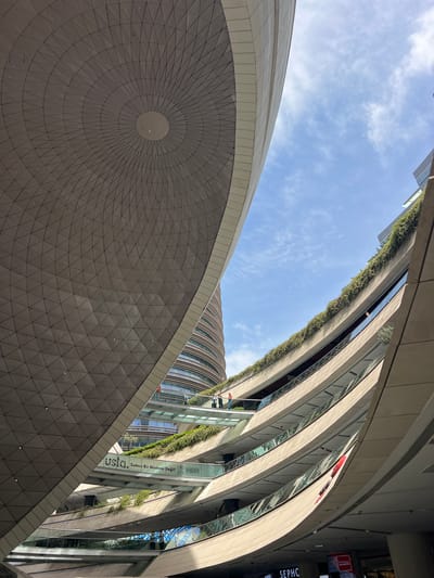 This is an eye-level, wide-angle shot looking up at the modern architecture of Kanyon Shopping Mall in the Şişli district of Istanbul, Turkey. The primary subjects are the layered, curved balconies and walkways of the mall, lush with greenery on the upper levels. A large, geodesic-like dome structure dominates the left side of the frame, its geometric pattern creating a visually striking contrast with the organic curves of the mall. In the background, a cylindrical tower building is visible. Several small figures, appearing to be people, are casually walking on a suspended glass walkway that connects different levels of the mall, offering a sense of scale to the vast structure. The sky above is partly cloudy with a clear blue expanse. The overall impression is one of contemporary urban design and a blend of nature and architecture.