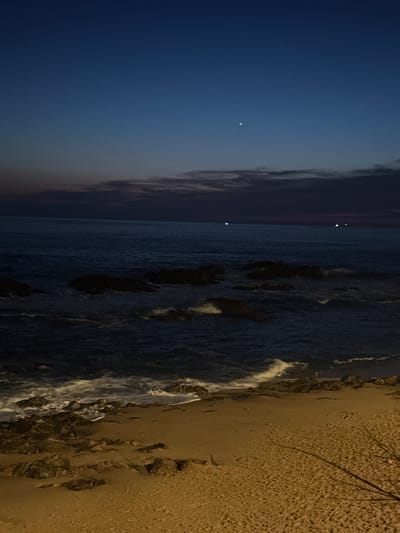 The image depicts a coastal scene at twilight or early night along the Atlantic coastline of Porto, Portugal, consistent with areas such as Foz do Douro or Matosinhos. The upper sky is a deep dark blue, gradually lightening to a purplish-grey horizon, with a bright celestial object positioned in the upper-middle left and dark, stratified clouds above the distant sea. The ocean appears dark blue-grey, featuring numerous dark, irregularly shaped rock formations partially submerged in the water. White-capped waves break against these rocks and advance onto the shore. Several small, bright white lights are visible on the distant ocean horizon, indicative of maritime traffic. The immediate foreground is a sandy beach, illuminated by an unseen artificial warm light source that casts an orange-yellow tone across the sand, where additional dark rocks are interspersed near the water's edge. No individuals or prominent anthropogenic structures are present in the observable scene.