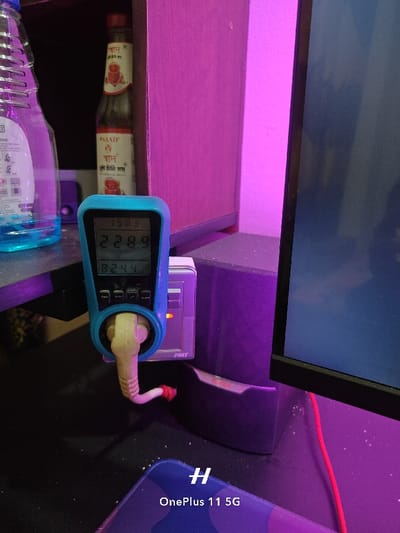 A blue digital power meter is plugged into an electrical outlet. The meter reads 228.9W and 824. The power meter is connected to a flat screen TV. The scene appears to be a bedroom or home office. The coordinates 24.388, 88.592 locate this image in Rajshahi, Bangladesh.