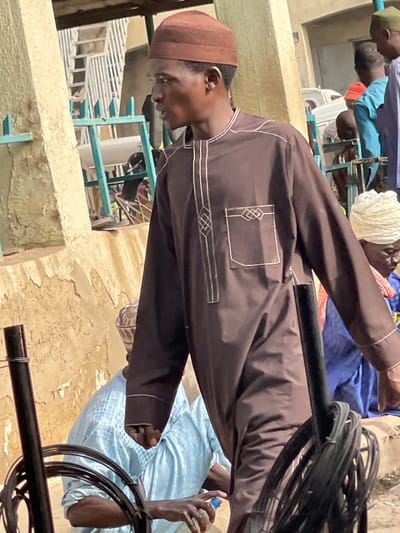 This image captures a group of men gathered in what appears to be an outdoor or semi-outdoor setting in Zaria, Nigeria, during bright daylight hours, suggesting sunny weather. The primary subject is a young man, centrally positioned and looking downwards, wearing a brown long-sleeved traditional garment, likely a thobe or kaftan, adorned with intricate white embroidery around a chest pocket and along the side. He also wears a textured brown kufi (prayer cap).
In the background, several other men are visible, some partially obscured. One man wears a light blue traditional garment, while others are seen in blue clothing, a white turban, and a green kufi, indicating a diverse array of attire common in the region. The men appear to be sitting or standing casually, possibly engaged in conversation or waiting, though no specific activity is clearly depicted.
The setting includes light blue metal structures, possibly chairs, tables, or part of a market stall, and concrete or mud-plastered walls and ground surfaces. A prominent black bar runs diagonally across the frame, with coiled black cables wrapped around one end, suggesting utility infrastructure. A hand holding a clear plastic bottle with a white cap is visible in the lower right, indicating the presence of refreshments. There is no discernible text visible in the image. The overall scene conveys a casual gathering in a functional, possibly community-oriented, environment.