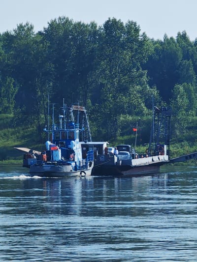 A tugboat named "DEM'SB" is pushing a barge across a wide, calm river. The tugboat, painted blue and white, has a Russian flag flying from its deck and several crew members in orange life vests visible. The barge, a flat-deck vessel, is carrying a dark-colored car and a light-colored SUV. A person in a white shirt stands near the SUV. The riverbanks are lined with dense green trees, characteristic of a forested area. The sky is clear and bright blue. The image captures a sunny day in the city of Galanino, Russia.