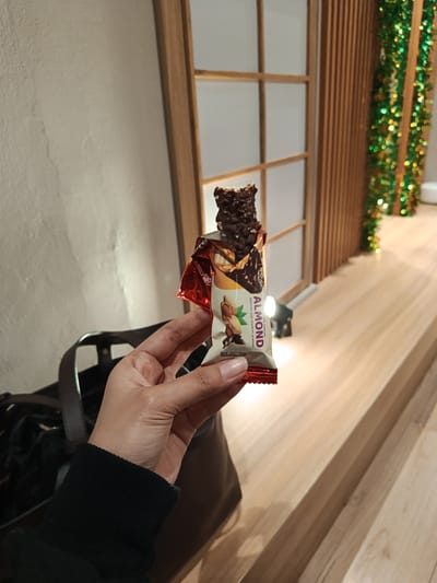 A person's hand holds a partially eaten chocolate bar indoors. The hand, with manicured nails, grips the colorful wrapper that features images of almonds and chocolate, and prominently displays text reading "ALMOND," "ROASTED AND CRUNCHY ALMOND," and "WHITE CARAMEL." An expiration date, "MAR 2027 FBH," is visible at the bottom left of the wrapper. The chocolate bar itself is dark brown and appears to be textured with visible pieces, possibly nuts or crisps.
In the blurred foreground, a dark-colored bag or purse is partially visible. The background consists of a light-colored wall on the left and a series of wooden lattice-like panels or screens on the right, suggesting a decorative interior window or partition. Below these wooden structures, a smooth wooden surface, resembling a shelf or counter, extends across the lower part of the frame. To the far right, a vertical strand of green and gold tinsel garland adds a festive touch to the setting. A small light fixture is visible on the wooden surface behind the chocolate bar. The scene is illuminated by artificial indoor lighting and is set in Bandung City, Indonesia, likely in a cafe, lounge, or home.