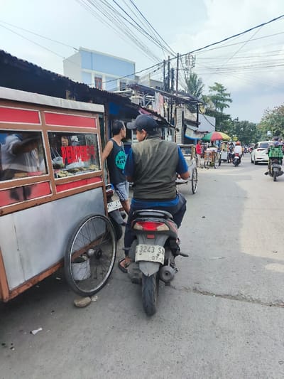 The image depicts a street scene in Bandung City, Indonesia, under a bright sky with dense overhead utility wiring. In the foreground on the left, a mobile food cart, red and metallic silver with glass display panels, is positioned on the asphalt pavement. The word "PEMPEK" is visible in red lettering on the cart, which contains yellowish food items. A person in a white shirt is partially visible inside the cart, attending to it. A detached bicycle wheel leans against the cart, propped by a rock, next to a small metal bucket.
To the right of the food cart, a man sits on a dark scooter, facing away from the camera, wearing a dark baseball cap, an olive green vest over a blue shirt, and dark pants with black sandals. The scooter's rear license plate, "D 3243 SJ 07.23," is clearly visible, identifying the vehicle as registered in Bandung. Another man stands to the left of the scooter, facing towards the food cart or the scooter rider, wearing a dark sleeveless shirt with white text and patterned shorts. A second scooter with license plate "D 341 FQ" is partially visible behind him.
The street extends into the midground and background, showing moderate traffic. Multiple motorcycles, some with delivery boxes, and at least one white car are present. Further down the street, a pedicab (becak) with an orange umbrella and other street vendors with carts are visible. Buildings line both sides of the street, including a prominent light blue and white structure in the background. Green foliage from trees is also discernible in the distance. The pavement is asphalt, showing signs of wear.