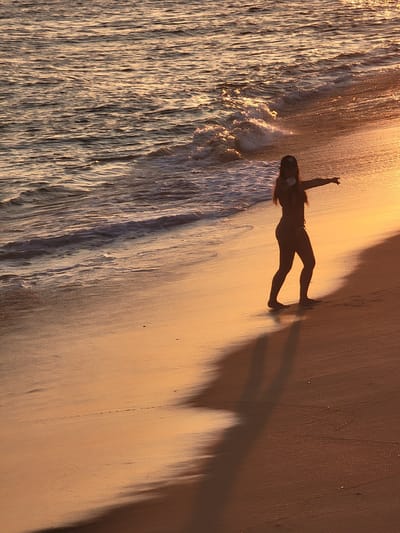 A silhouette of a person in a bikini and cap stands on a sandy beach at sunset, with their right arm extended towards the water. The person is positioned to the right of the frame, with their shadow cast on the wet sand. The ocean, with gentle waves and reflections of the setting sun, occupies the upper left portion of the image. The warm, golden light of the sunset illuminates the beach and the water. The scene appears to be on a beach in Quarteira, Portugal.