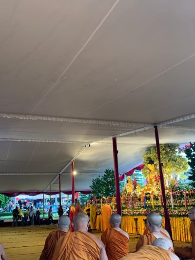 The image shows a group of Buddhist monks gathered under a large white tent in Mungkid, Indonesia. Located at coordinates (latitude: -7.605, longitude: 110.23), the scene is likely part of a religious ceremony. The monks, wearing orange robes, are facing a golden statue of Buddha decorated with flowers and candles, positioned on an ornate altar at the back of the tent. The monks are bowing their heads in prayer, and a few people are standing behind them observing the ceremony. The tent is decorated with white fabric and has several red poles supporting it. Outside, a few trees and a green field can be seen. The overall atmosphere is calm and spiritual.