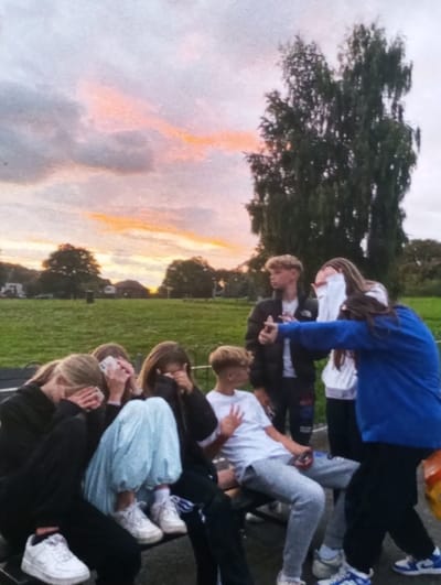 The image displays an outdoor scene at twilight, consistent with a park or recreational area in Brzezina, Poland. A group of seven young individuals, appearing to be teenagers, occupies the central and foreground areas.

In the foreground, a dark metal bench is visible. Three individuals are seated on the left side of this bench, with their faces obscured by their hands or personal items. One individual wears dark clothing and white sneakers. Next to them, another wears a dark top, light blue baggy pants, and white sneakers. The third wears dark clothing.

To the right of these individuals, a male with blond hair, wearing a white t-shirt and grey sweatpants, sits on the same bench, making a two-finger hand gesture with his left hand. Behind him, another male, wearing a dark jacket and dark pants, stands holding a dark, indistinct object in his right hand.

To the right of the male in the dark jacket, a female in a white hoodie and dark pants is partially visible. Further to the right, another female, dressed in a bright blue long-sleeved top and dark pants, stands with her right arm extended towards the male in the dark jacket. All individuals are wearing casual attire.

The ground immediately surrounding the bench appears to be dark, possibly paved or gravel. The background consists of a large, green grassy field with several trees. A prominent, tall, slender tree with dark foliage stands on the far right. Distant, indistinct buildings are faintly visible on the horizon. The sky exhibits patches of orange and pink coloration from the setting sun, contrasted against a predominantly cloudy, light grey backdrop.