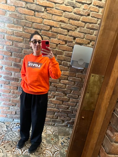 An indoor scene shows a female subject taking a mirror selfie. The subject, an adult female with dark hair pulled back, wears dark-framed eyeglasses, an orange crewneck sweatshirt with a reversed white "LEVI'S" logo, black wide-leg pants, and dark footwear. She holds a red smartphone in her right hand, displaying light blue nail polish. The background behind the subject and reflected in the mirror consists of an exposed brick wall of varying orange and brown tones. The floor features patterned tiles in a mosaic of colors including brown, beige, blue, and white. To the right, within the mirror's reflection, a light gray wall-mounted dispenser is visible, alongside a dark wooden frame with a brass-colored metallic accent. The image contains no discernible identifiers for the city of Argveta, Georgia.