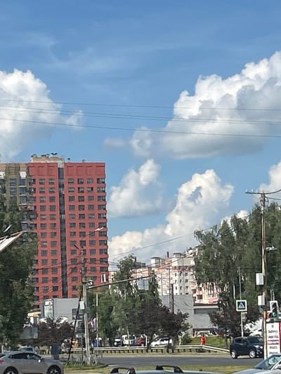 This media file depicts a street scene in Zhukovo, Russia, on a bright, sunny day with scattered clouds. The perspective is from a vehicle traveling along a road. Dominating the foreground is a tall, reddish-brown brick building with numerous windows, suggesting it's an apartment complex or office building. To the right, a road is visible with several cars, including a silver sedan in the lower right corner. Trees line the side of the road, providing a green contrast to the urban architecture. In the background, other buildings and more trees are present. Overhead, power lines crisscross the sky, and fluffy white clouds drift against a clear blue sky, indicating fair weather. A traffic light with green and red signals is visible further up the road, along with a pedestrian crossing sign and a traffic sign with a white diamond shape and a blue border. The scene suggests a typical day in a populated area, with ongoing traffic and everyday urban life. There are no visible people clearly identifiable in the scene. The overall impression is one of a vibrant, active daytime environment.
