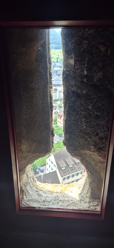 This is a vertical, eye-level, point-of-view shot taken from inside what appears to be a stone structure, looking through a narrow opening. The opening frames a view of the town of Sargans, Switzerland. The foreground is dominated by the rough, textured stone of the window embrasure, which is illuminated by sunlight. Beyond the stone opening, a town landscape unfolds. Buildings with pitched roofs, some with visible windows and architectural details, are clustered together. In the middle ground, a modern white building with green shutters stands out. Further back, a wider vista shows rolling green hills, with trees and fields, and in the distance, mountains under a bright sky. A small dark car is parked on a street in front of one of the buildings. The overall impression is of an ancient perspective overlooking a contemporary European town and its surrounding natural beauty.