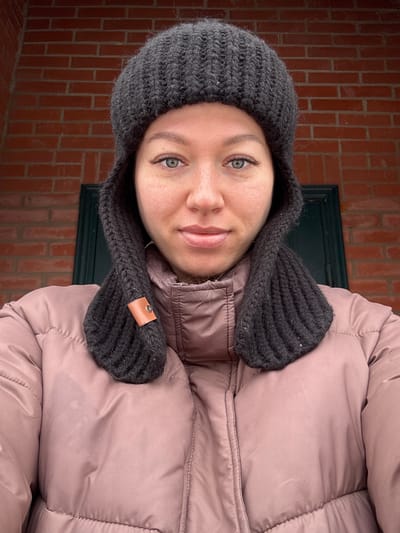 A woman is standing in front of a red brick wall, likely an exterior building facade. She is wearing a rose-colored puffer jacket and a dark grey knit hat with ear flaps. A dark green window or door frame is visible behind her. The woman's face is centered in the frame; she is looking directly at the camera with a slight smile.
The location mentioned, Ферма, Russia, cannot be verified from visual information alone.