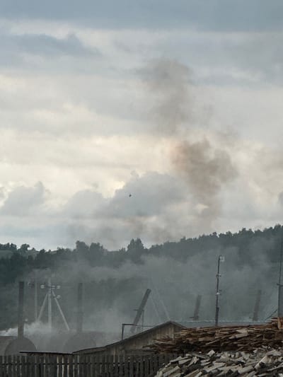 A wide shot captures a landscape dominated by a cloudy, overcast sky. In the middle ground, a dense line of trees forms a dark silhouette against the lighter sky. Below the tree line, a hazy layer of smoke or fog obscures industrial structures. These structures appear to be cylindrical, with tall, slender poles or chimneys rising from them. Some of these poles are topped with lights. The foreground features a collection of lumber, piled high in disorganized stacks. A wooden fence runs across the bottom of the image, partially obscuring the view of the lumber and industrial area. The overall impression is one of an industrial site or lumber processing facility, possibly in a rural or forested area, with the weather contributing to a somewhat muted and hazy atmosphere. The location is identified as Abzakovo, Russia.