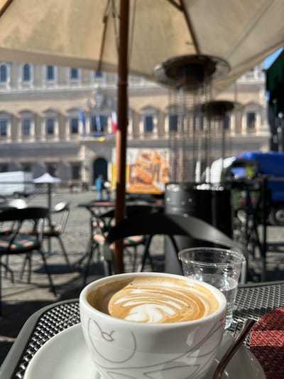 A cup of cappuccino sits on a table at an outdoor cafe in front of Palazzo Montecitorio in Rome, Italy (41.895, 12.471). The cappuccino has a distinct latte art design. The table is a black metal table with a black mesh top. The cafe has black metal chairs, a white umbrella and several outdoor heaters. In the distance, several people can be seen walking on the cobblestone street. In the background, Palazzo Montecitorio is visible. The building has a white facade and is adorned with flags. A blue truck is parked to the right of the cafe, partially obscured by the outdoor heaters. The sun is shining brightly. The scene is typical of an outdoor cafe in Rome, where people come to enjoy a leisurely drink and take in the sights of the city.