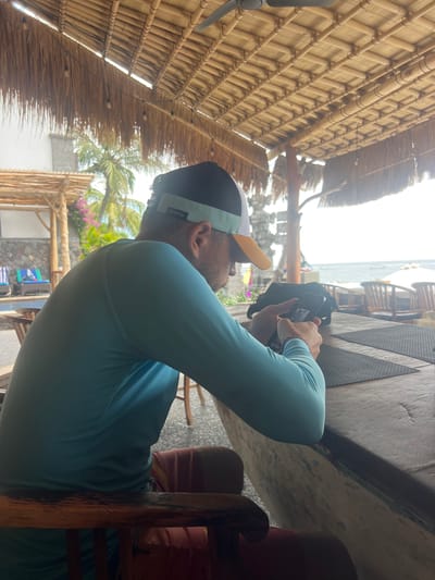 A man wearing a light blue long-sleeved shirt and a cap sits at a wooden table under a thatched roof. He is looking down at a mobile phone in his hands. In the background, palm trees and the ocean are visible. The man is seated at a bar or restaurant, possibly in Abang, Indonesia, given the tropical setting and architecture. The area appears to be a resort or beachside establishment.