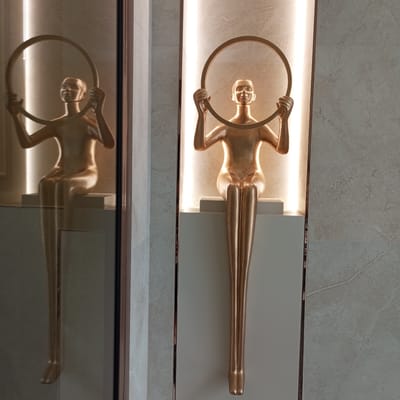 This image shows a golden statue of a person holding a hoop, displayed in a well-lit alcove. The statue is seated on a white platform, with its legs elongated and streamlined. The background consists of marble tiles and a vertical strip of bright light. To the left of the statue, a reflective surface creates a mirrored image of the sculpture. The scene appears to be an indoor installation, possibly in a public space or a modern residential building. There is no indication of the specific location within the city of Pervomaysky, Russia, as the image does not contain any recognizable landmarks or identifying features of the city.