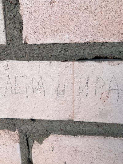 This media file shows a close-up, eye-level view of a brick wall. The bricks are a light, speckled beige color, and they are joined together by dark gray mortar. The mortar has an uneven, rough texture, with some areas appearing chipped or broken away, revealing the underlying materials.

There is writing scratched into the surface of two of the bricks. On the top brick, the writing appears to be in cursive and is partially obscured. The visible letters suggest the word "Mila" or "Vila." On the brick below, there are more clearly discernible letters scratched into the surface. The top portion of this writing appears to be "M" and "I", followed by what could be "L" or "V", and then "A". The writing is in capital letters and is arranged vertically. The overall impression is that of graffiti or casual etching on the brick surface.

The lighting suggests it is daytime, with no strong shadows indicating a particular time of day. The setting appears to be an outdoor environment, given the texture of the bricks and mortar, and the likely outdoor nature of graffiti. There are no people or other objects visible, focusing the viewer's attention solely on the wall and its markings. The context provided indicates the location is Sinyavino, Russia.