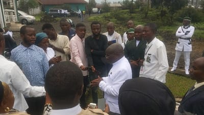 The image shows a group of people gathered in front of a building. The building appears to be made of concrete and has a green door. There are trees and foliage in the background. The people are all dressed in formal attire, including white shirts, suits, and ties. The man in the white shirt and glasses is holding a smartphone and is looking at it. He is surrounded by other men in suits. The man in white and a green cap is standing to the right, facing the camera. There are two men in military-style uniforms in the background. One of the men in military uniform has his arms crossed, and the other is standing at attention. The people are in Keshero, Democratic Republic of the Congo, at the coordinates -1.623 latitude and 29.157 longitude. They are likely gathered for a formal event.