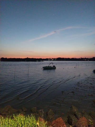 The image shows a coastal scene at dusk in what appears to be Badem, India. The sky exhibits a color gradient, transitioning from a deep blue on the left to an orange-pink hue along the horizon, suggesting either sunrise or sunset. A strip of land is visible along the horizon, sparsely dotted with lights. In the water, a small, dark green boat is anchored. Wooden stakes are placed in the water. The water is slightly rippled. The right edge of the frame is a shoreline with dark, muddy soil and some green vegetation. A rocky border partially separates the water from the foliage. The image is oriented sideways, with the sky on the left and the water on the right.